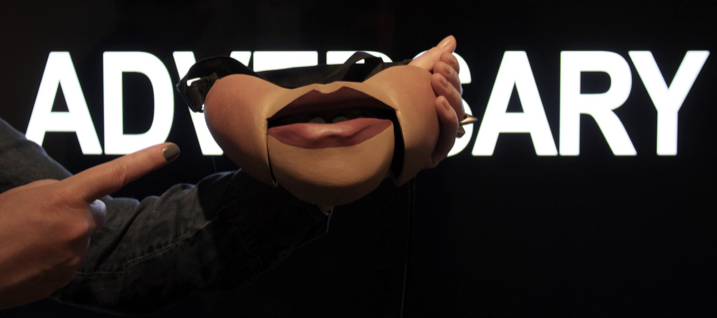 image of ventriloquists half mask (mouth) with the word 'adversary' in the background