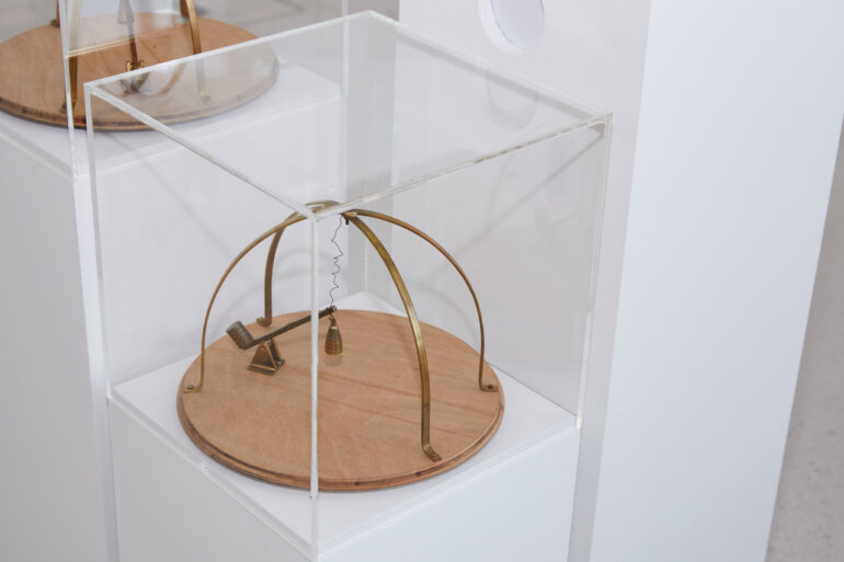 kinetic artwork of a circular wooden base and brass arches with a small brass seasaw with a weight. on a white plinth under a clear acrylic box