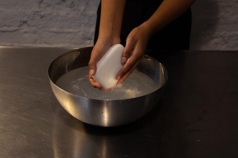 Close-up of a pair of hands holding a bar of ivory coloured soap in a silver coloured metal bowl of water.